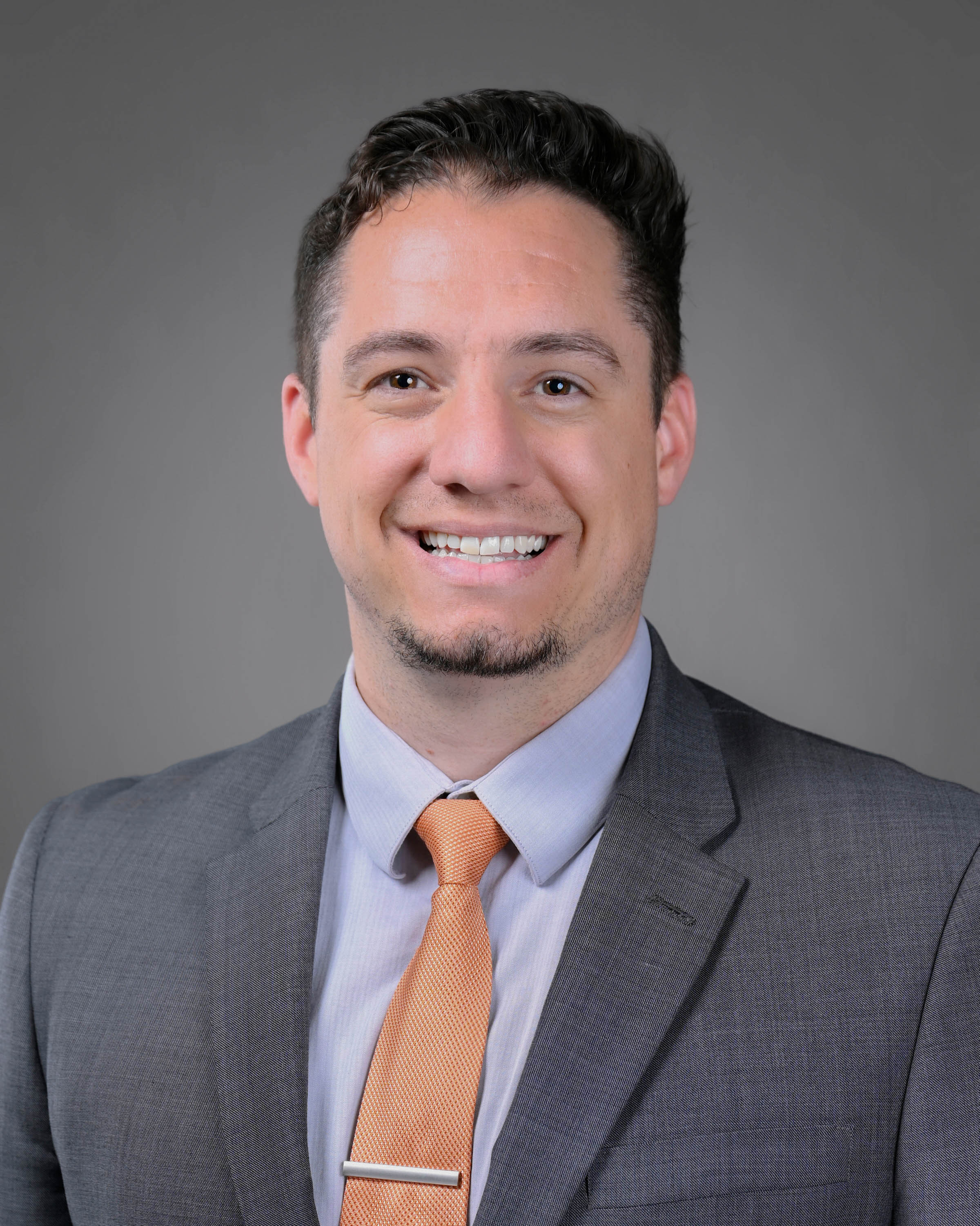 Man smling with short dark brown hair wearing a grey jacket and orange tie